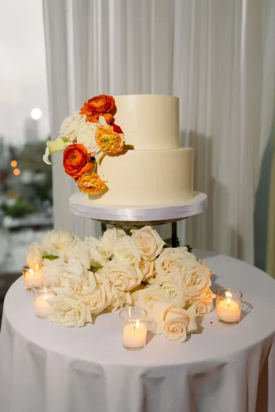 Wedding cake, a two-tier cake glowing beside votive candles, set before sheer drapes and a window with a cityscape backdrop.