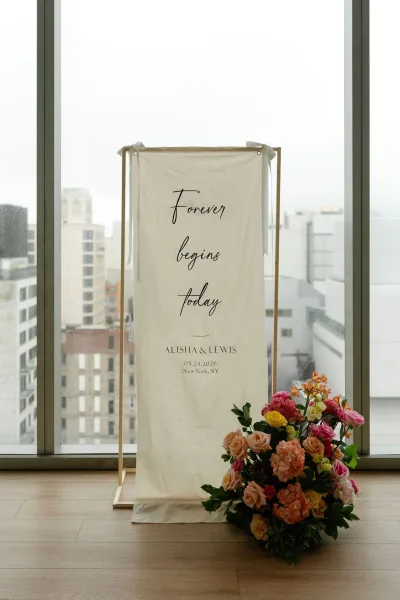 Wedding welcome sign on a fabric banner sign, accented by a floral arrangement, set before large windows overlooking city buildings.