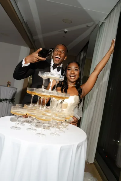 Champagne tower moment with a wedding champagne pour cascading into coupe glasses as joyful newlyweds celebrate in an indoor venue with curtains.