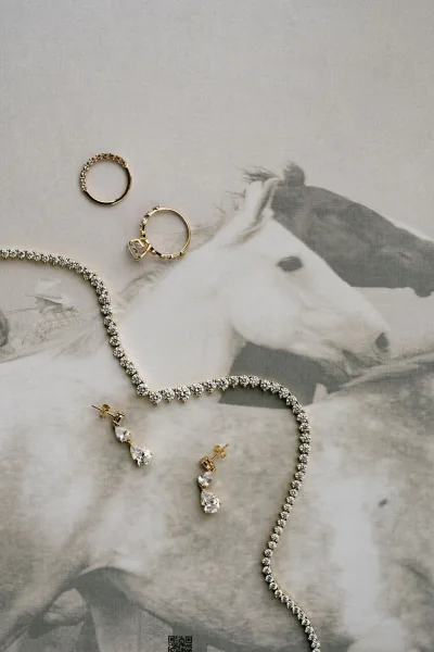Bridal jewelry styled as an engagement ring flatlay with a sparkling tennis necklace on fabric beside horse print artwork.