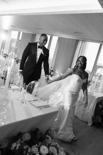 Couple portrait of a bride and groom holding hands, her veil visible as they stand by windows with a city view.
