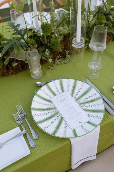 Reception tablescape with green wedding tablescape details, patterned plate and menu card, white napkin, silver flatware, ribbed glasses, tall candles, and lush greenery centerpiece