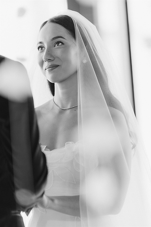 Bridal portrait of a bride wearing veil, smiling up at the groom’s shoulder in soft window light, sheer tulle veil framing her face