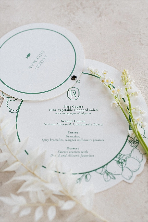 Wedding menu card with scalloped edge and green ink typography, monogram crest and grommet tag beside lily of the valley on stone surface