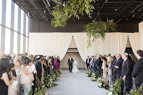 Wedding processional with bride walking down aisle on a white runner, groom waiting near draped curtains and hanging greenery in a bright indoor venue