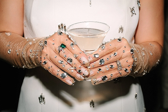 Bridal cocktail detail with bride holding martini, crystal mesh gloves and jeweled gown, engagement ring and band against dark backdrop