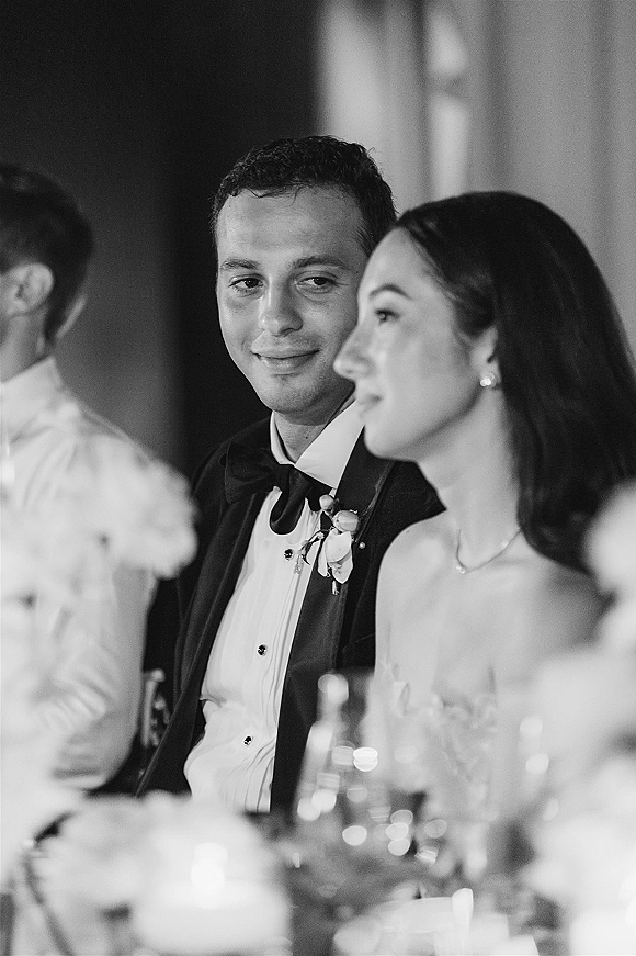 Wedding reception moment as bride and groom at table share a candid toast, tuxedo bow tie and wine glasses at the head table with guests behind