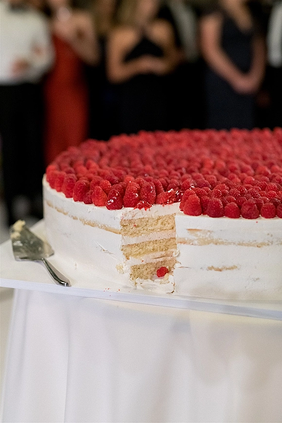 Wedding cake with raspberries on white frosting, a slice missing and cake server on stand as guests mingle in the reception space