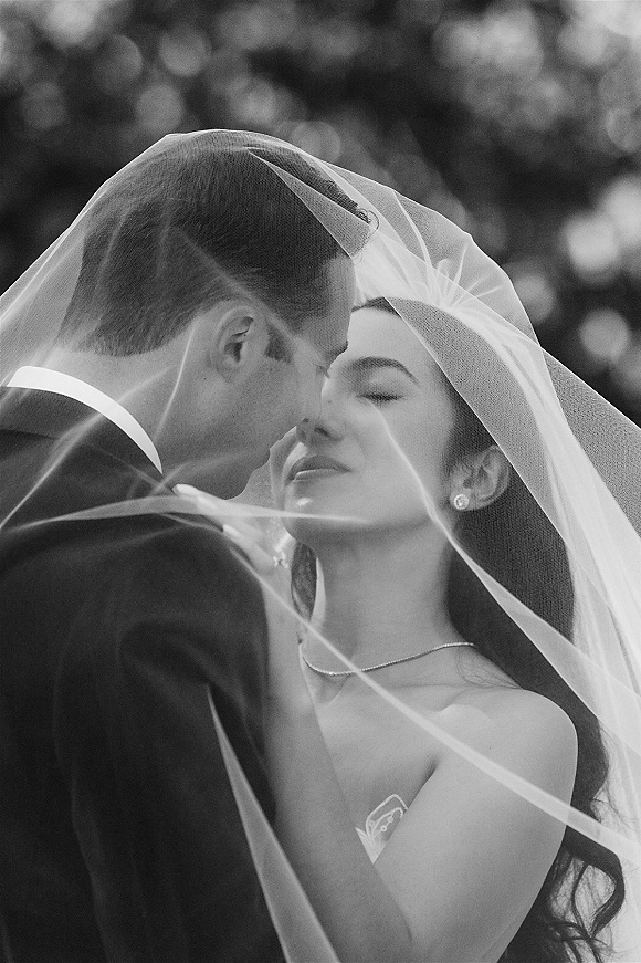 Wedding couple portrait in black and white, bride and groom under veil touching foreheads, bride holds his face with trees softly blurred behind