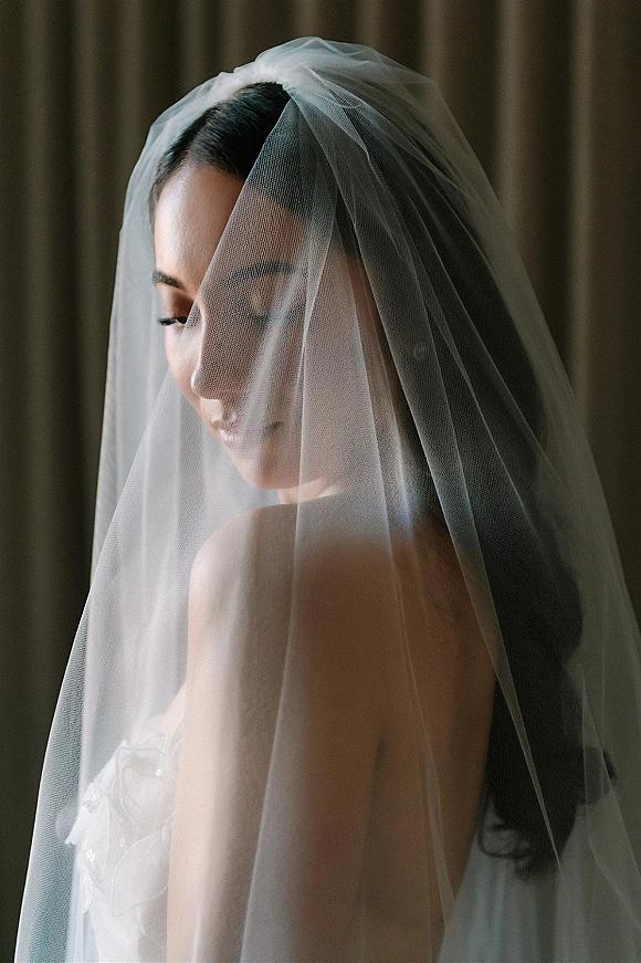 Bridal portrait of a bride with veil draped over her face, wearing a strapless wedding dress and earrings against softly lit curtains indoors