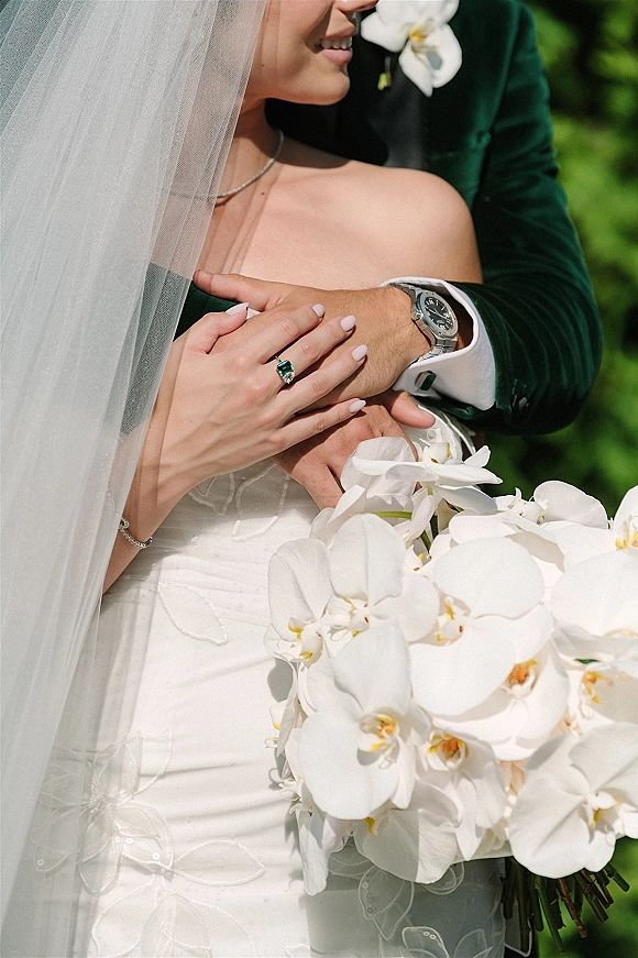 Couple portrait of bride and groom embrace, bride in veil holding cascading orchid bouquet with emerald ring against outdoor greenery