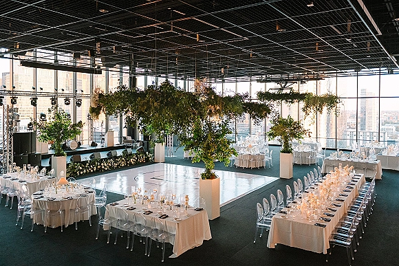 Reception decor with a modern wedding reception look, featuring a hanging greenery installation over candlelit tables and a white dance floor by city-view windows