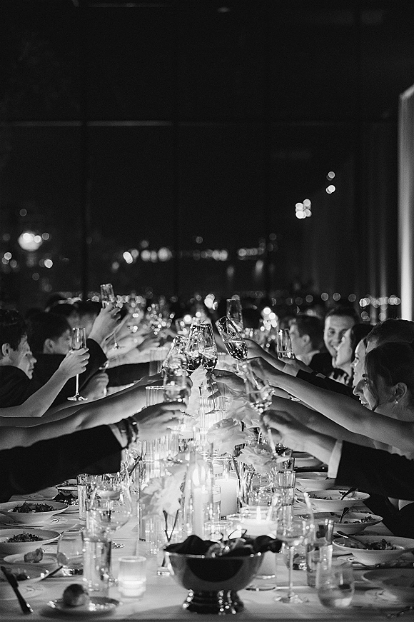 Wedding reception toast as guests clink champagne flutes over a candlelit banquet table, with city lights glowing beyond the window wall at night