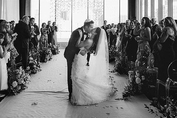 Wedding kiss as newlyweds share a first kiss, bride in cathedral veil and groom in tuxedo on floral aisle with city skyline windows
