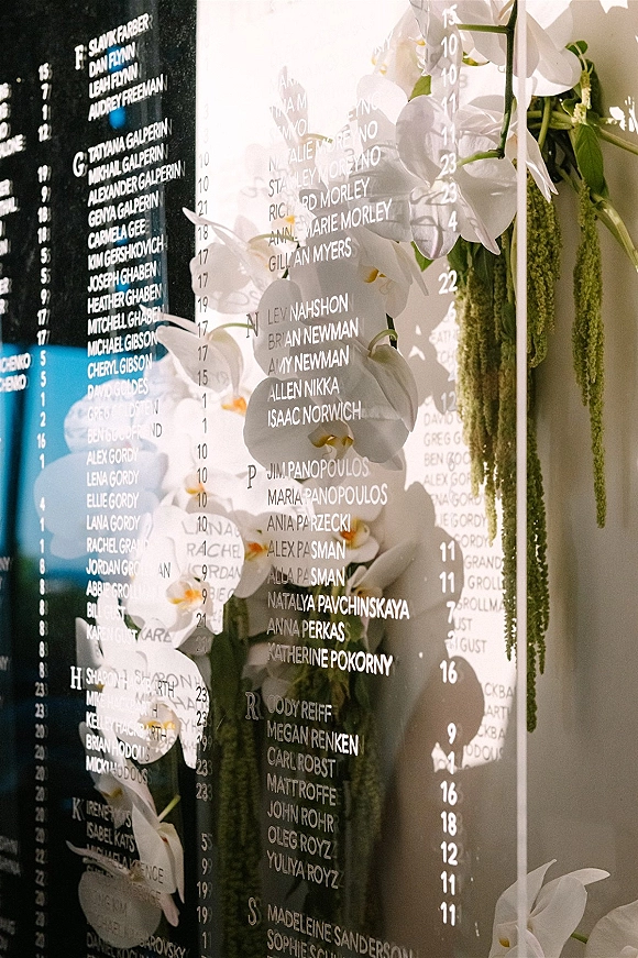 Wedding seating chart on acrylic seating chart with printed guest names, white orchids and hanging greenery against a neutral wall with black directory board
