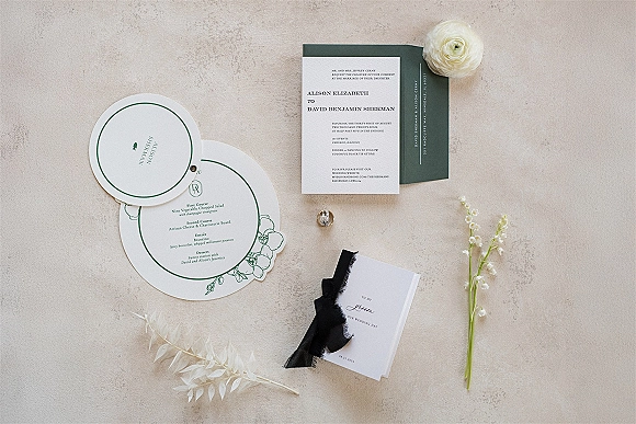 Invitation suite with modern wedding invitations, black ribbon and wax seal beside a white rose on a neutral stone surface flat lay