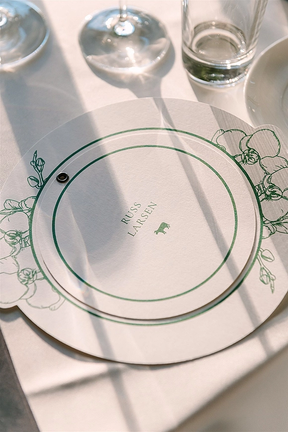 Wedding place card with a round wedding place card design, green illustration and name typography on a plate beside glasses on linen in sunlight shadows