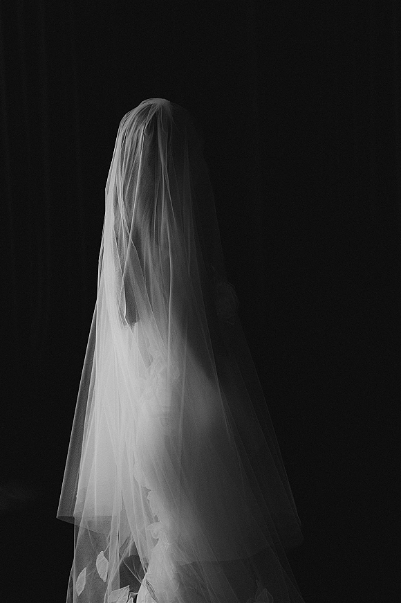 Bridal portrait in black and white of a bride in veil, her sheer wedding veil and gown softly lit against a dark studio backdrop