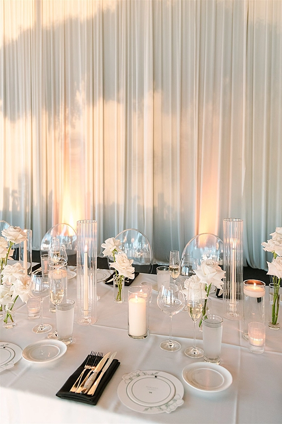 Reception tablescape with white rose centerpieces and a candlelit wedding table set with gold flatware, black napkins, and clear acrylic chairs against a white draped, uplit backdrop