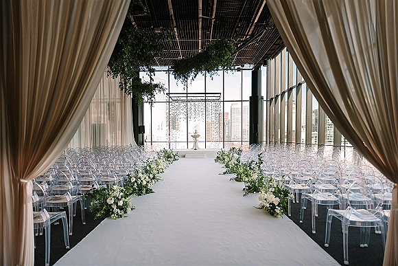 Ceremony setup for an indoor wedding ceremony with a white aisle runner, clear acrylic chairs, and greenery by city-view windows