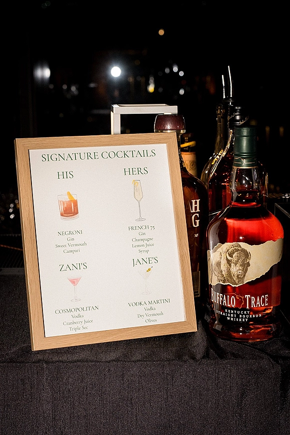 Signature cocktails sign in a framed display on a wedding bar with liquor bottles and pour spouts, set against dark bokeh lights