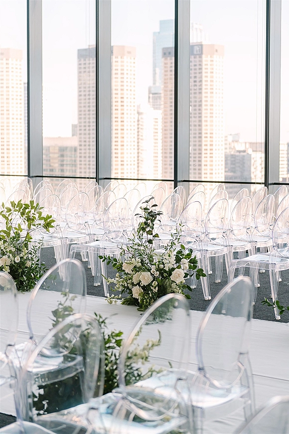Ceremony setup with clear acrylic wedding chairs lining a white aisle runner, framed by white florals and greenery before city skyline windows