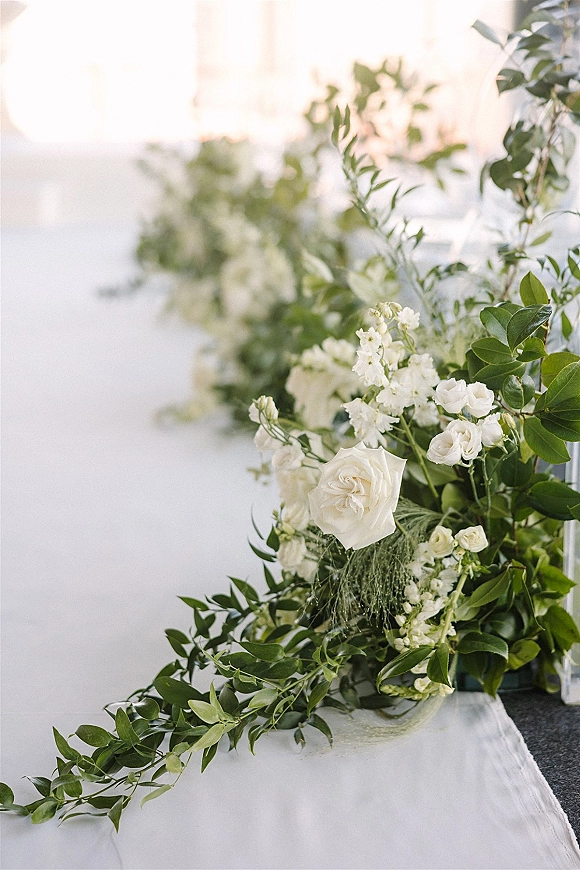 Wedding floral garland with a greenery table garland of white roses and foliage laid on a white tablecloth in bright window light