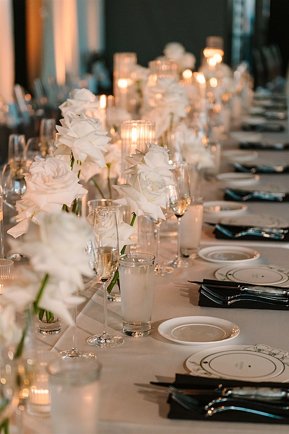 Reception tablescape with a white rose centerpiece, taper and votive candles, and black napkins on a long banquet table indoors