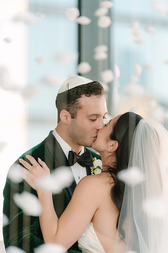 Wedding kiss portrait of bride and groom kissing, her veil draped over strapless dress, by glass windows with soft floral foreground