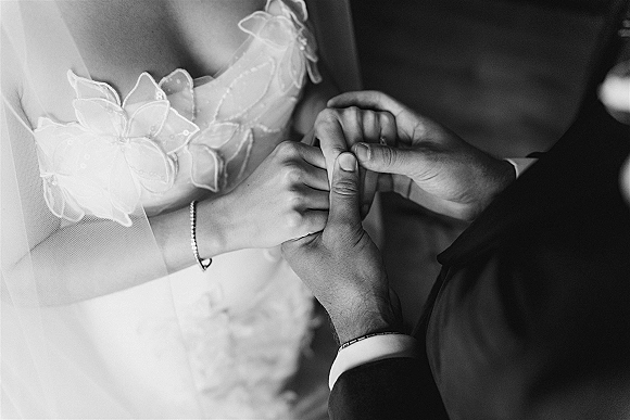Wedding ring exchange as groom slides a band onto the bride’s finger, her lace bodice and veil visible against a blurred indoor backdrop
