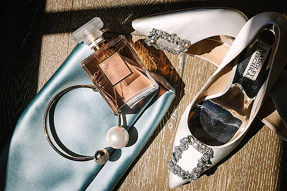 Bridal accessories flatlay with white satin heels and rhinestone buckles beside perfume, silk clutch, and pearl bangles on sunlit wood floor