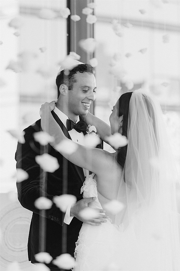 First dance as bride in veil and strapless gown embraces groom in tuxedo under hanging flowers in a window-lit reception space