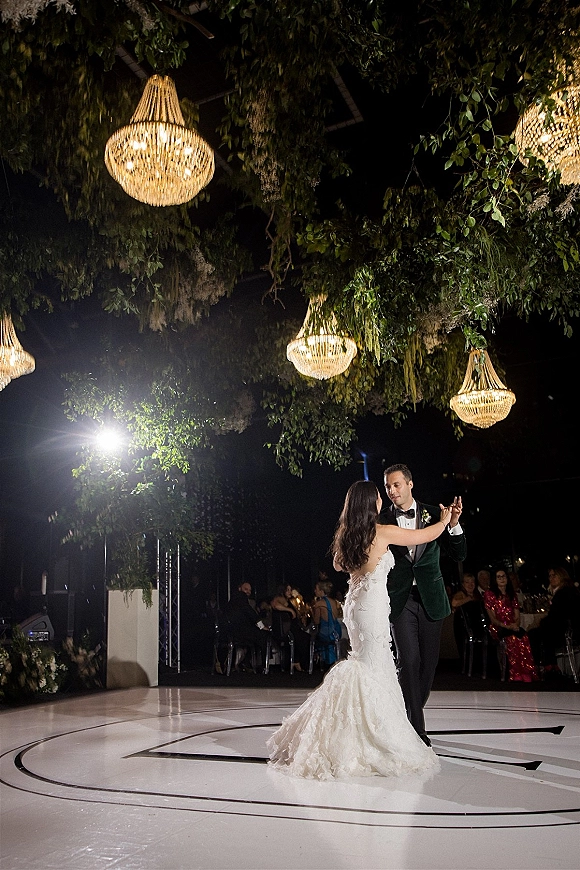 First dance as bride and groom dancing on a monogrammed floor beneath chandeliers and greenery, with guests at tables under night sky