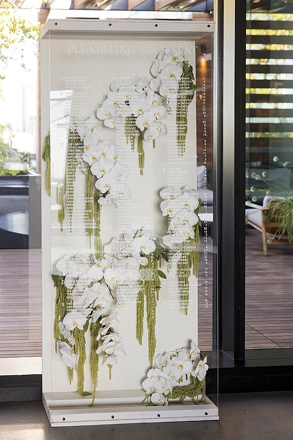 Seating chart display on a clear acrylic seating chart board with printed guest list text, framed by white orchids and hanging greenery by glass doors