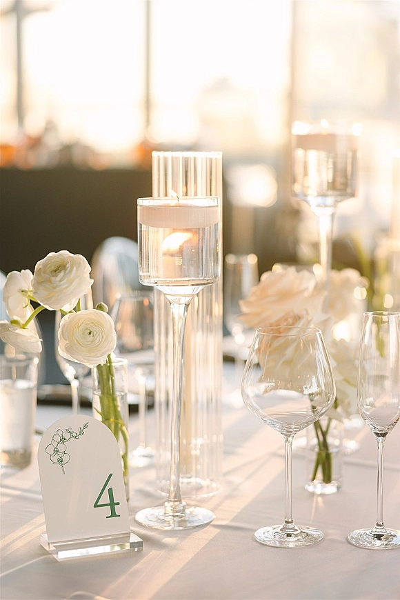 Reception tablescape with floating candle centerpiece in glass cylinder vases, blush and white flowers, and clear acrylic table number in window light