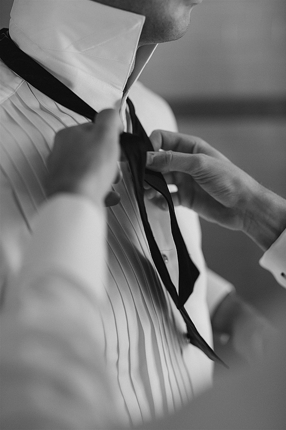 Groom getting ready adjusts his groom bow tie over a pleated tuxedo shirt with wing collar and jacket cuff in a softly blurred room