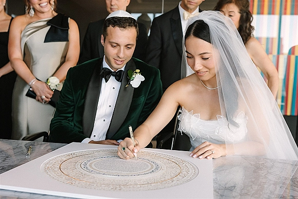 Wedding signing as bride in strapless dress and veil signs marriage certificate on marble table while groom in velvet tux watches indoors