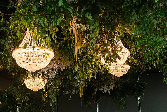 Reception ceiling decor with a greenery ceiling installation, crystal chandeliers and hanging foliage garlands with baby’s breath on a dark ceiling