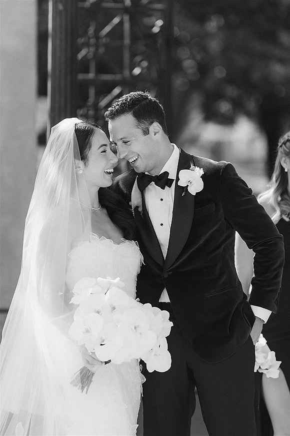 Couple portrait in a black and white wedding portrait, bride and groom laughing as she holds an orchid bouquet on a columned walkway