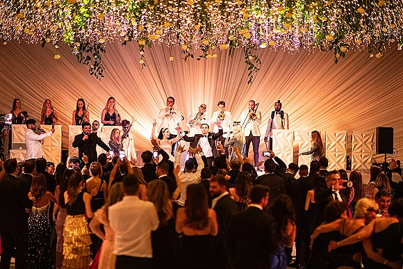 Wedding reception party with a wedding band performance on stage, brass instruments and string lights above a packed dance floor crowd indoors