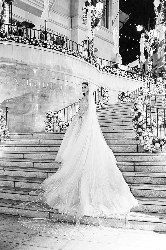 Bridal portrait of a bride on staircase, looking back with a long veil and sweeping dress train amid garlands and string lights