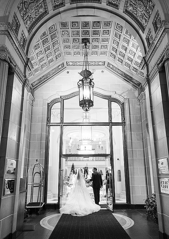 Wedding exit of bride and groom leaving venue, seen from behind with long veil and gown train under ornate arched lobby entryway