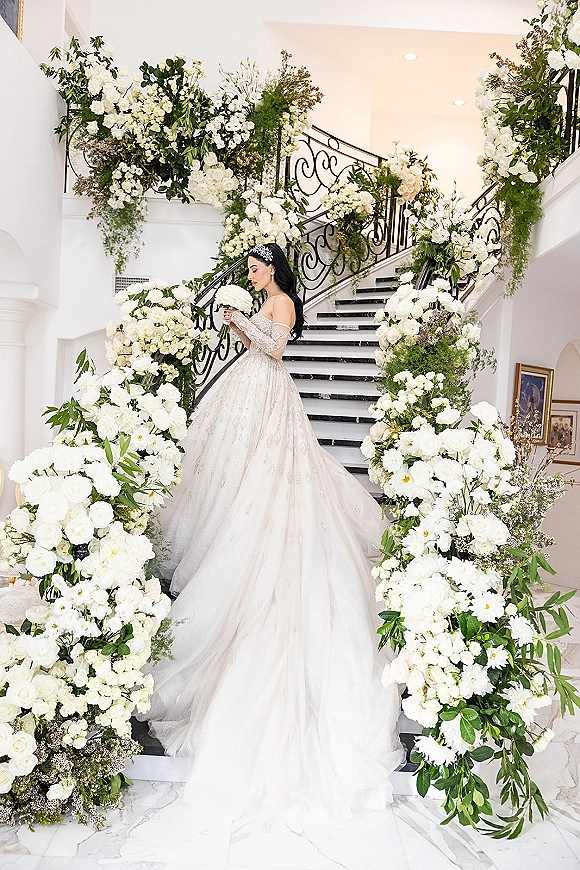 Bridal portrait of a bride holding bouquet in a sparkly ball gown and long veil on a floral-adorned indoor staircase with wrought iron railing