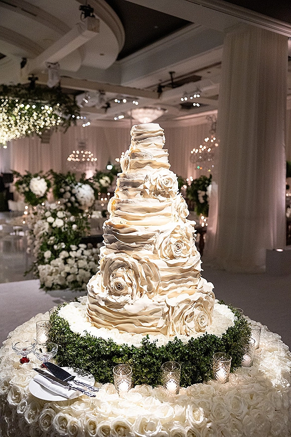 Wedding cake with tiered wedding cake ruffled buttercream and white rose accents, surrounded by votive candles under chandeliers and draped curtains