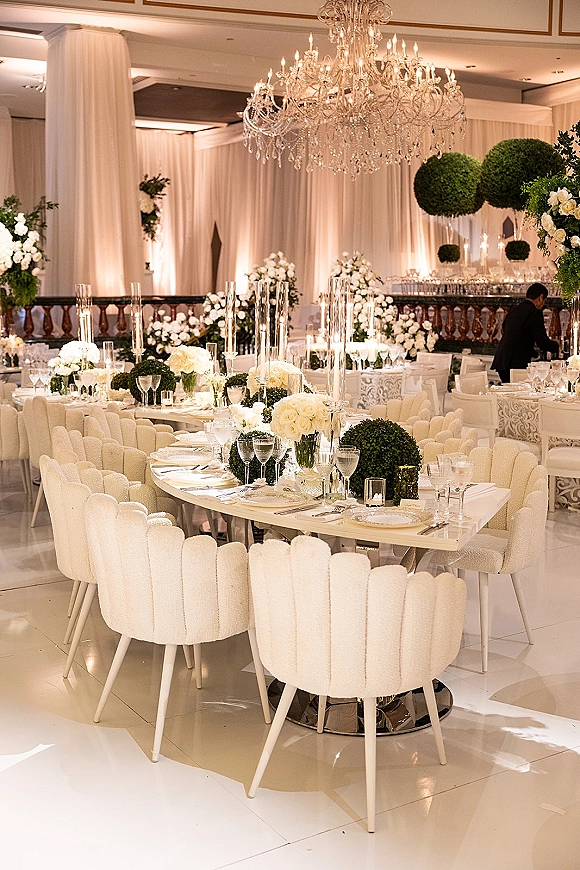 Reception tablescape at a luxury wedding reception with white rose centerpieces, candles in glass cylinders, and a crystal chandelier in a ballroom