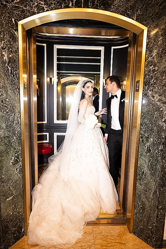 Couple portrait in an elevator wedding photo, bride in strapless gown with cathedral veil and bouquet beside groom in tuxedo by gold doors