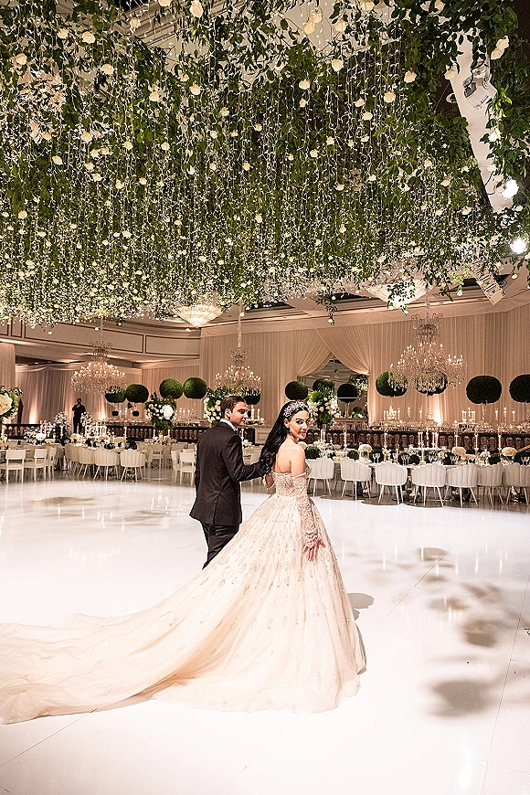Couple portrait of bride and groom holding hands on a white dance floor under hanging greenery, chandeliers, and candlelit tables