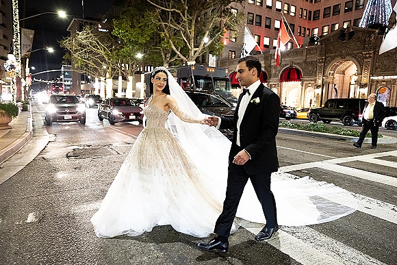 Couple portrait of bride and groom walking hand in hand across a city crosswalk at night, her long veil trailing under streetlights