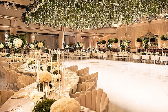 Reception decor with white floral centerpieces, candlelit tables, and hanging greenery ceiling beneath chandeliers in an elegant ballroom