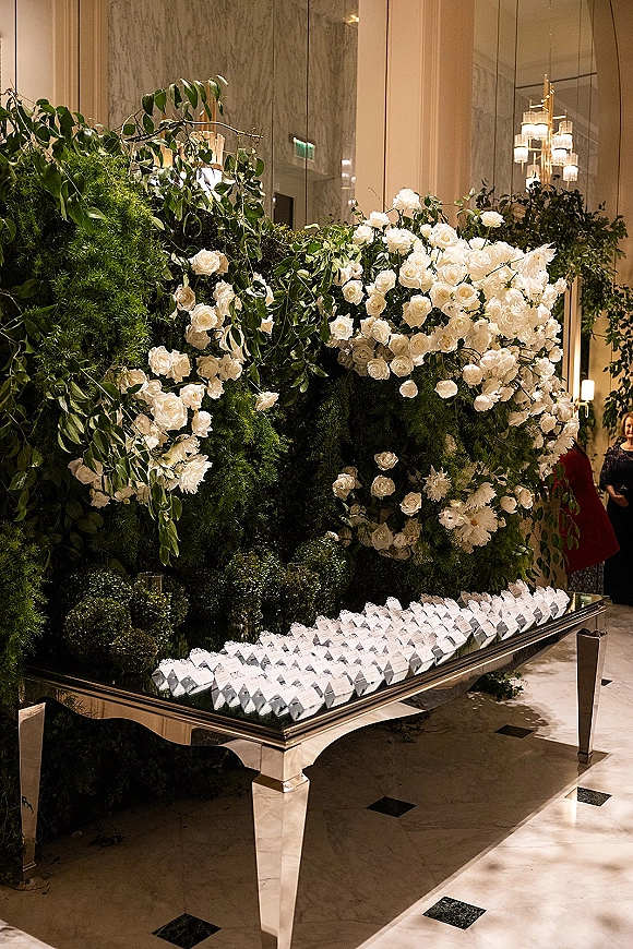 Wedding escort card display with escort card table ideas on a mirrored table, white roses and greenery wall, under chandeliers in a marble hall